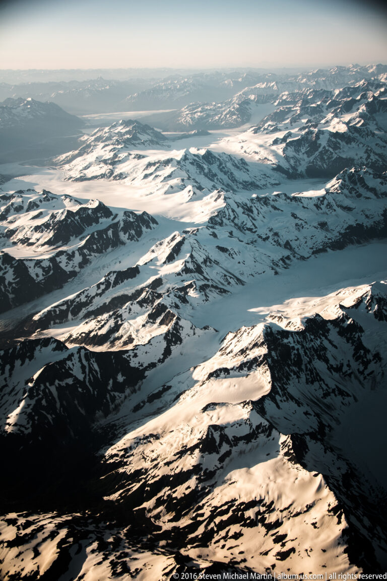 Sunlit snowfields and glacier channels extend across rugged mountains toward a pale horizon.