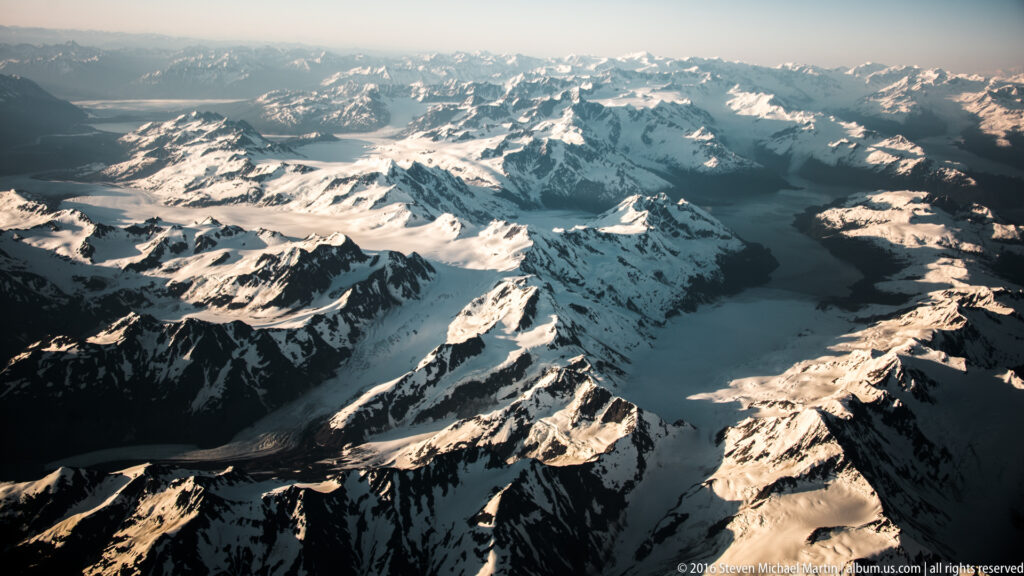 Jagged snowy peaks rise above narrow glaciers and shadowed mountain valleys beneath warm light and a hazy sky.
