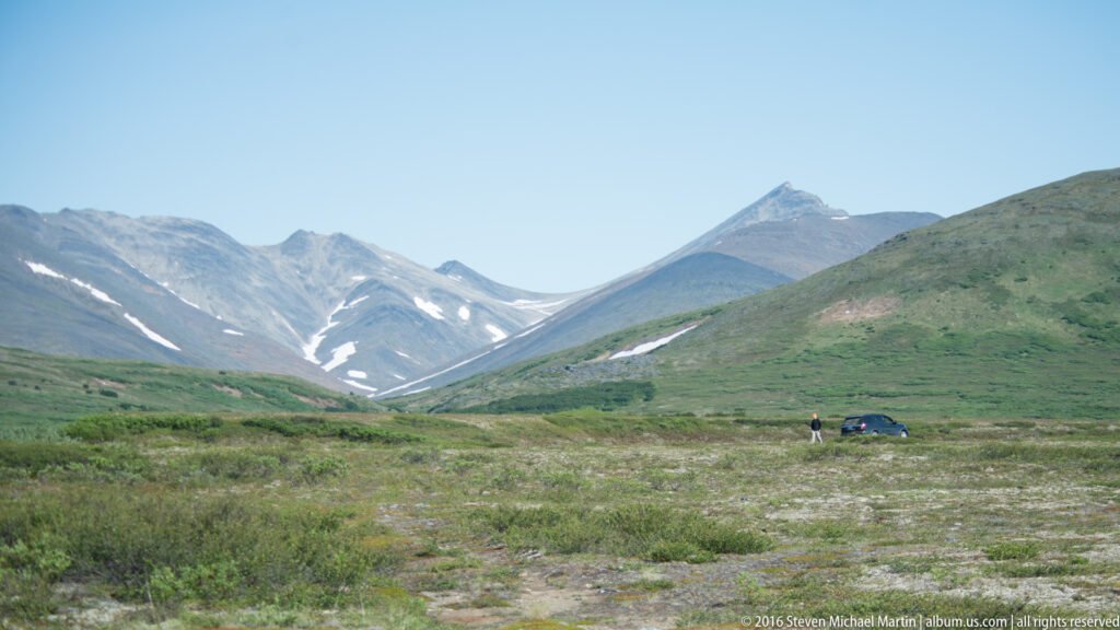 A person and vehicle stand in a broad tundra valley below snow-streaked mountains.