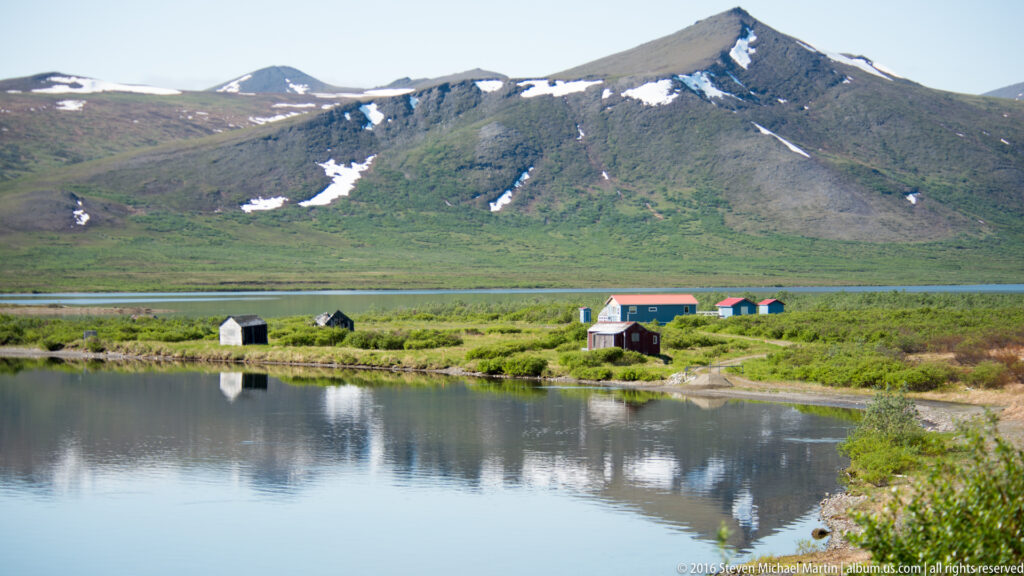 Small red and blue buildings sit between two lakes beneath a snow-patched mountain.