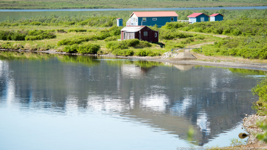 Small lakeside buildings and sheds reflect in calm water along a remote shore.