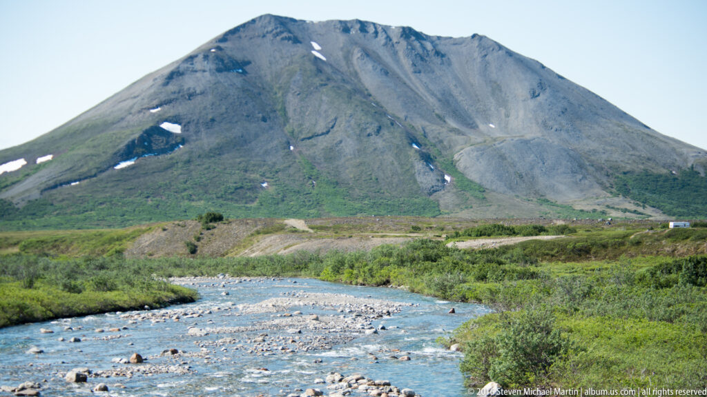 A rocky river flows beneath a steep snow-patched mountain and green tundra slopes.