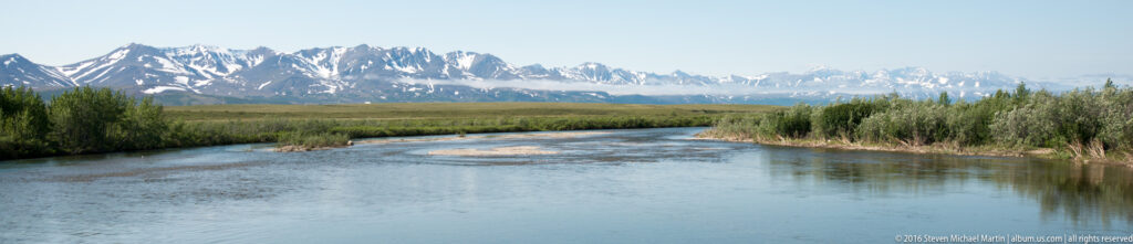 A river curves through a forested foreground toward distant snow-covered mountains in Alaska.