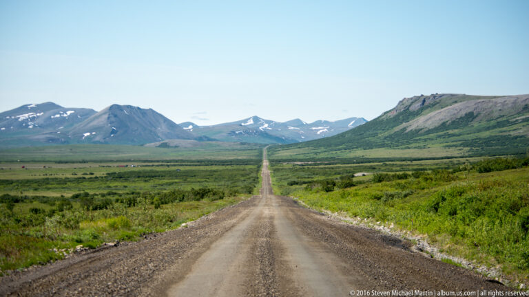 A gravel road leads straight through a broad tundra valley toward distant mountains.
