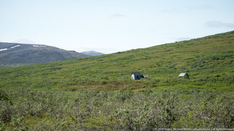 Two small cabins stand far apart on a broad green tundra hillside.