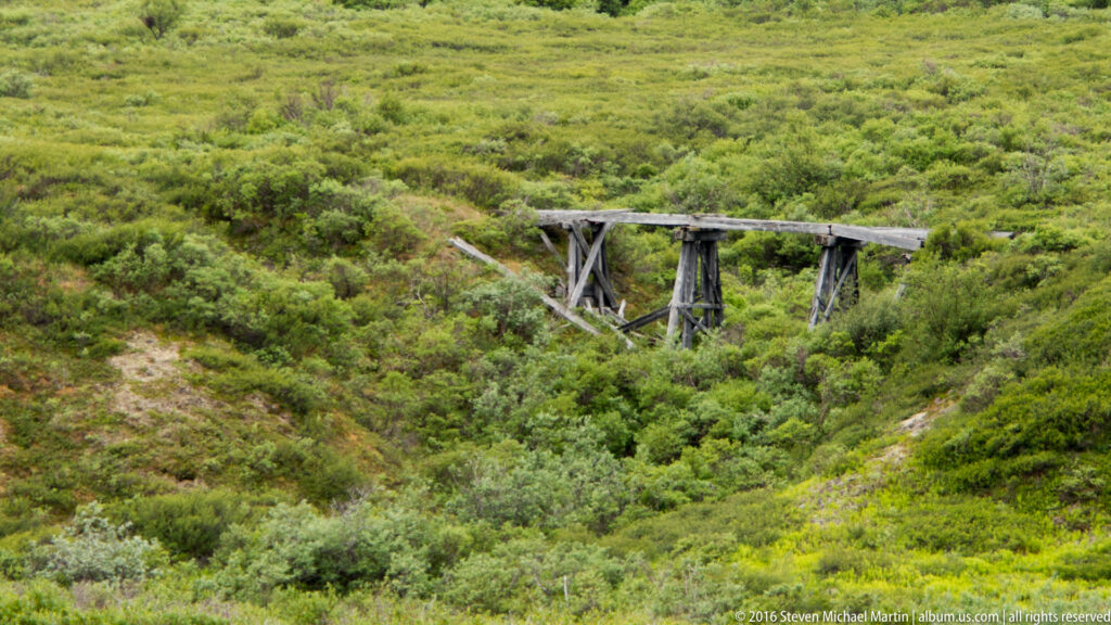 A weathered timber trestle bridge crosses a shrub-filled ravine in green tundra.