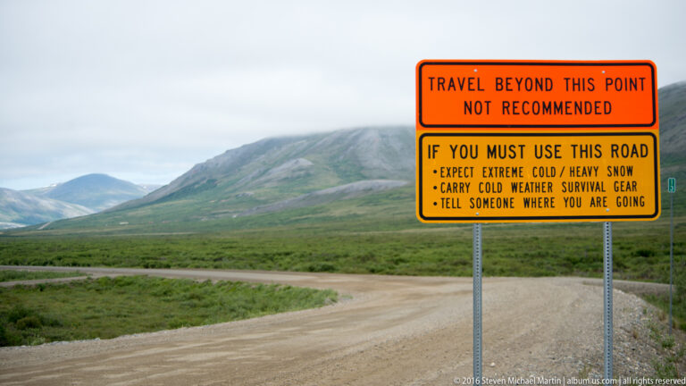 A roadside warning sign stands beside a gravel road in a remote mountain valley.