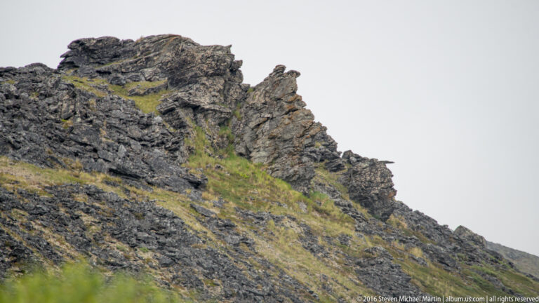 Dark fractured rock rises diagonally above a green tundra slope beneath pale sky.