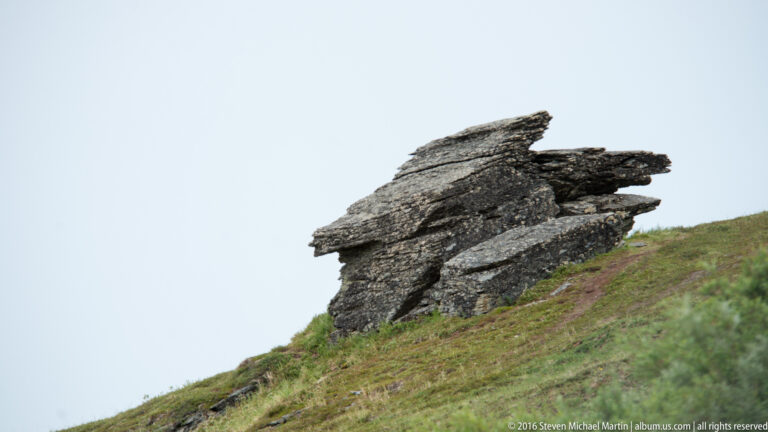 A tilted, layered rock formation projects from a grassy hillside against open sky.