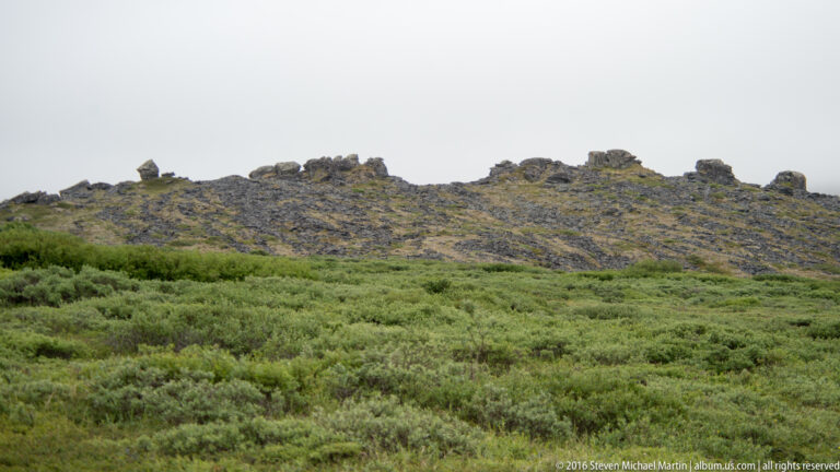 A broken line of dark rocks crosses a green tundra hillside beneath a pale sky.