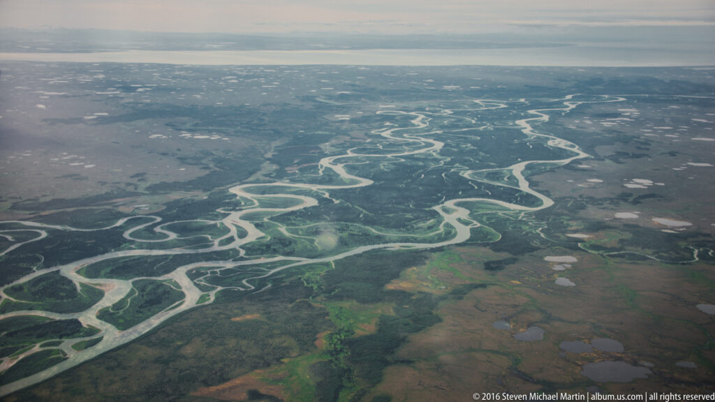 Aerial view of pale braided river channels winding through flat green-brown wetlands toward the horizon.