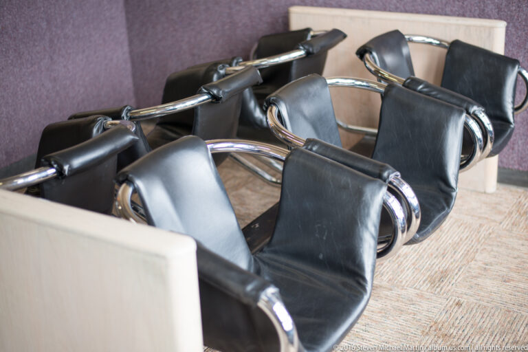Rows of black airport waiting chairs with chrome frames beside a pale partition and muted carpet.