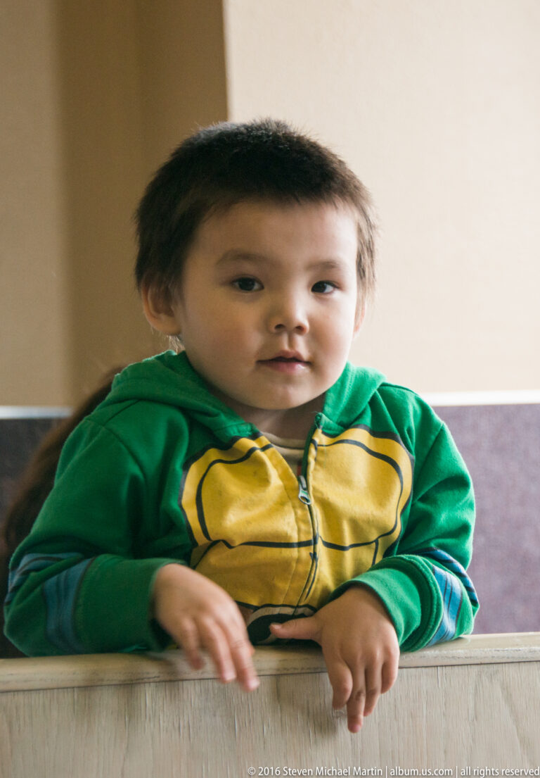 Young child in a green hoodie leaning both hands on a pale counter inside a softly lit room.