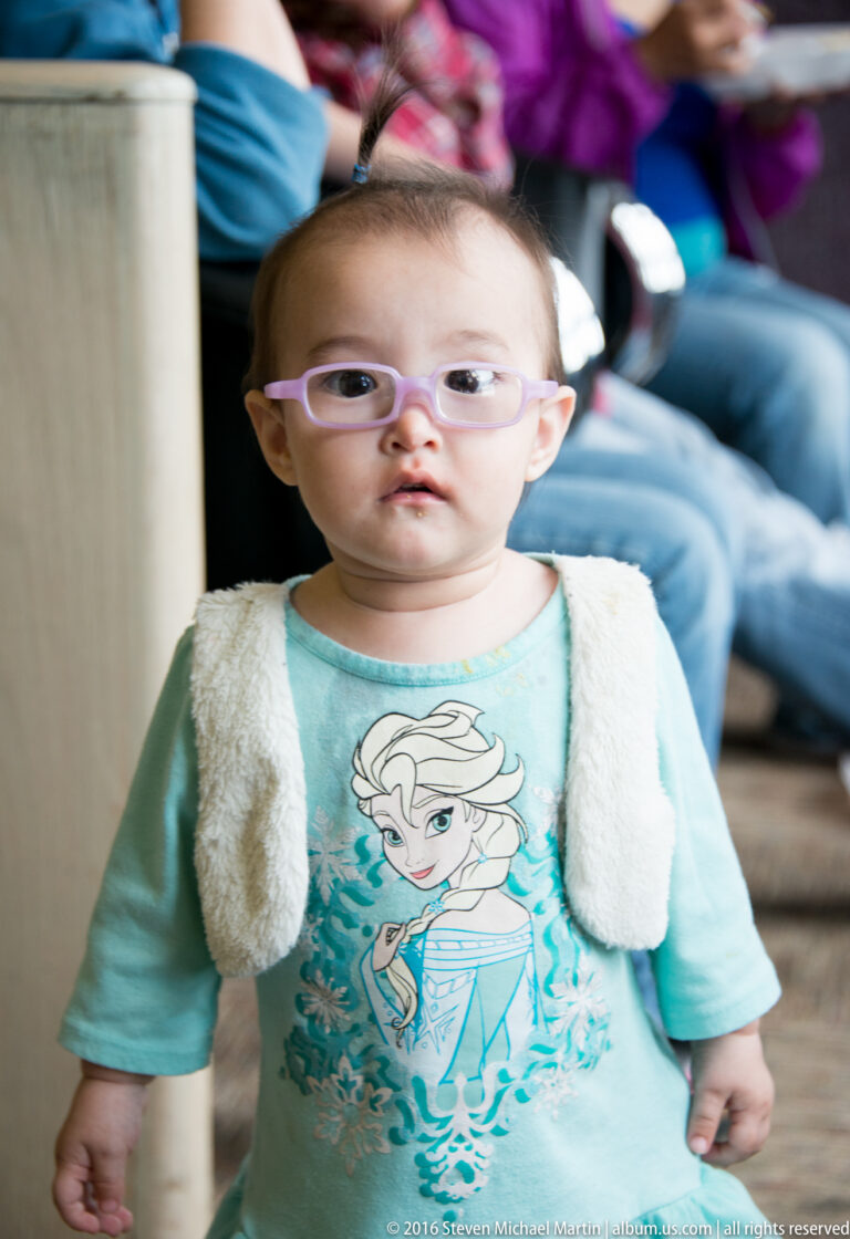 Young child in lavender glasses and a pale vest standing before a softly blurred group of seated people.
