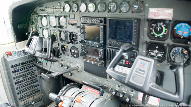 Aircraft cockpit with two control yokes, instrument gauges, radios, switches, and a central display.