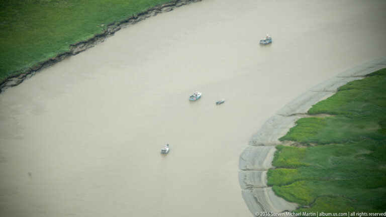 Several small boats on a wide tan river beside a narrow strip of green shoreline.
