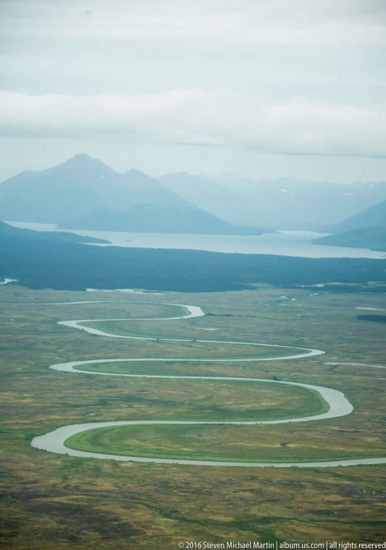 Wide river loops across flat tundra toward distant blue water and low mountains beneath an overcast sky.