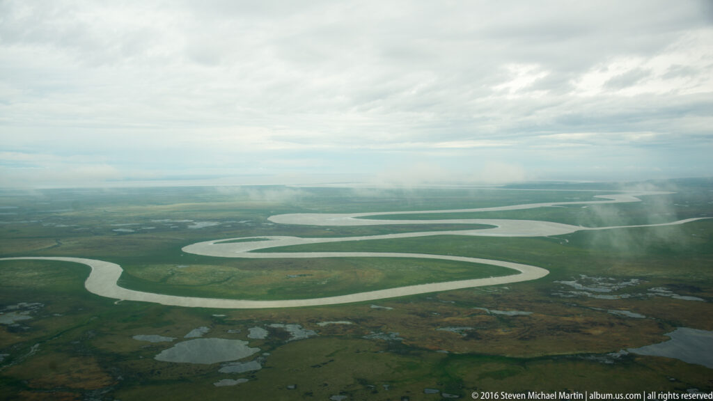 Pale river bends through green wetlands beneath low cloud, with small ponds scattered across the plain.