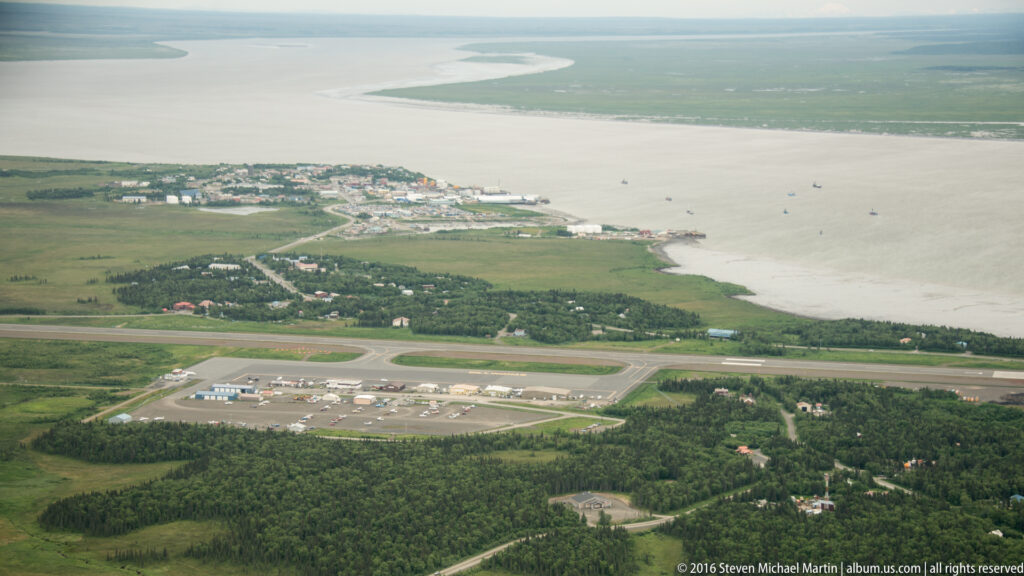 Aerial view of Dillingham, with its runway and buildings between green terrain and a broad river mouth.