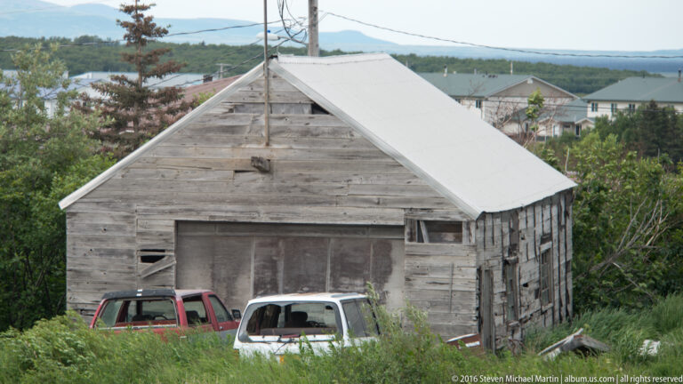 Weathered gray wooden shed with a steep gable roof, partly screened by shrubs and parked vehicles.