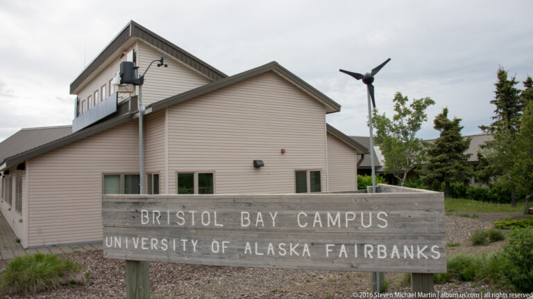 Wooden sign for the Bristol Bay Campus, University of Alaska Fairbanks, in front of a low building and small wind turbine.