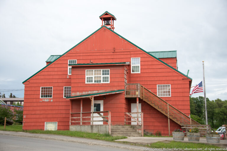 Large red gabled building with white-trimmed windows, a small cupola, exterior stairs, and an American flag.