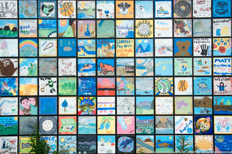A large rectangular grid of colorful small paintings and drawings covers an outdoor wall.