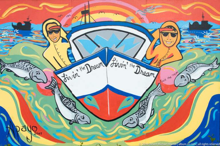 Colorful painting of two people in a boat labeled “Livin’ the Dream,” surrounded by salmon in swirling blue water.