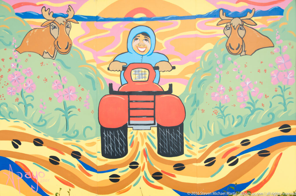 A child rides an orange all-terrain vehicle toward the viewer, with two moose in a colorful painted landscape.