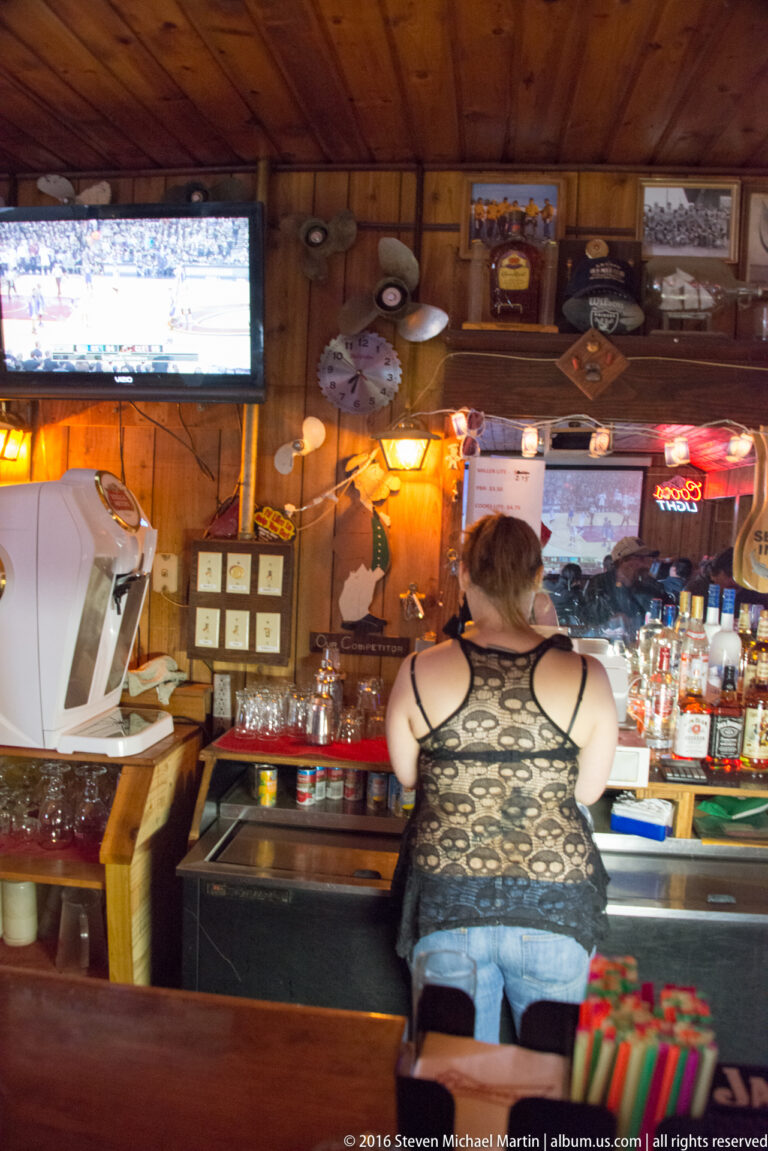 A customer stands at a counter in a wood-paneled bar lined with bottles, framed memorabilia, and a television.