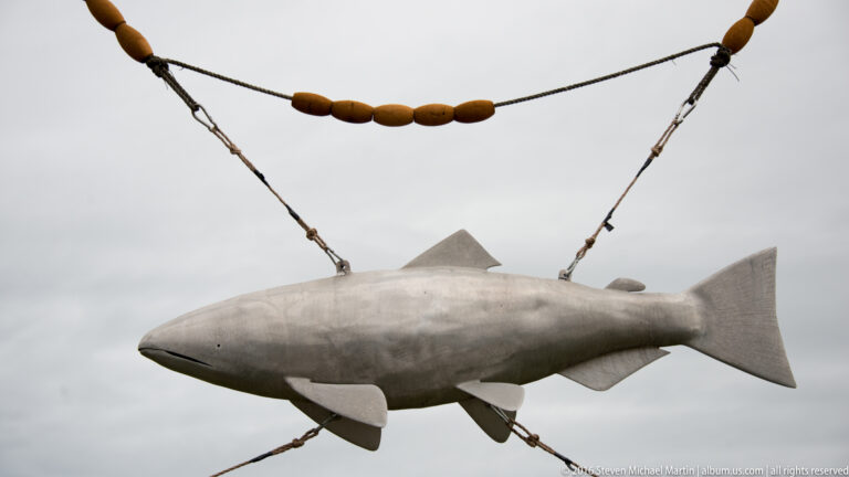 A large gray fish sculpture hangs from cables, with brown oval elements strung along the line above it.