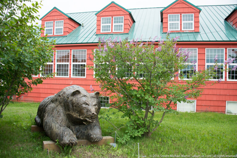 A carved bear sculpture on a low plinth sits in a lawn before a coral-red building with dormer windows.