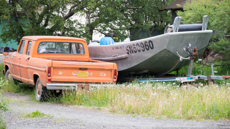 An orange pickup is parked beside an aluminum boat on a trailer in a grassy yard.