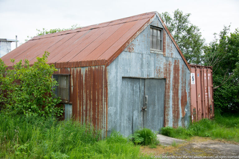 Rust-streaked corrugated-metal shed with a reddish roof stands in grass, with a shipping container behind it.