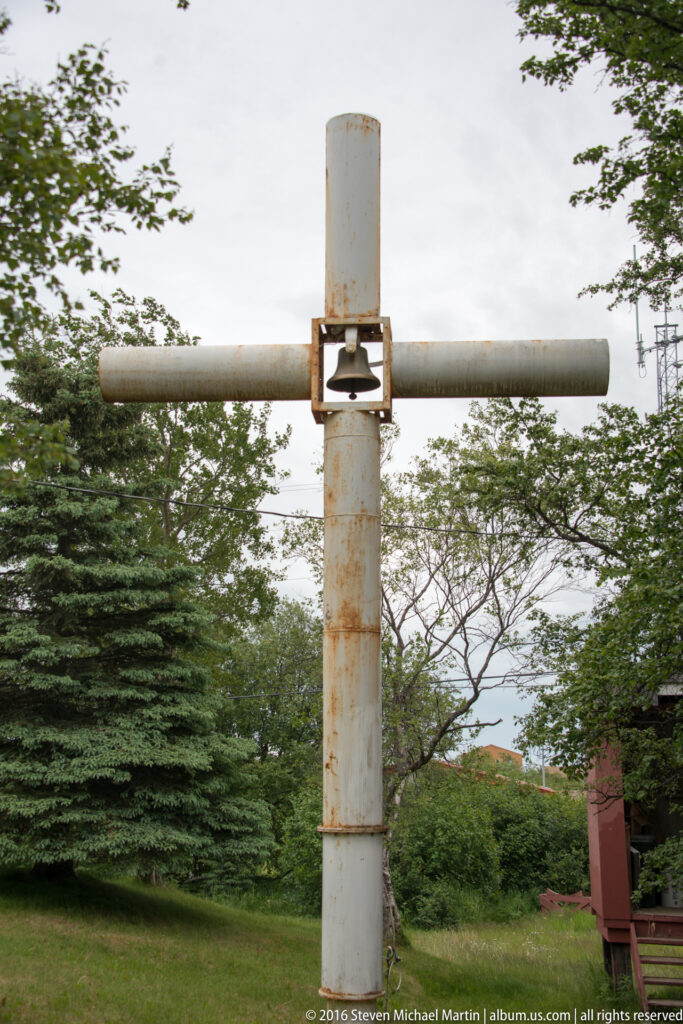 Rusted vertical metal pole with a horizontal crossbar and central fitting stands among trees beside a red utility box.
