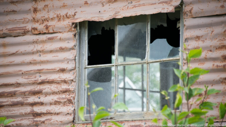 Broken upper panes in a weathered multi-pane window set within peeling pale clapboard siding.