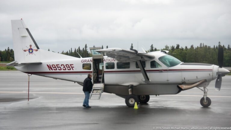 White single-engine airplane with an open cabin door and boarding steps sits on wet pavement beside a person.