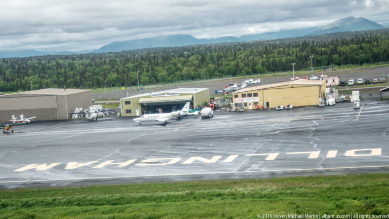 Airport apron with small parked aircraft and low service buildings, backed by forested hills under clouds.