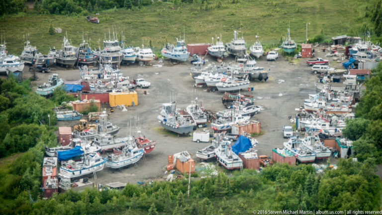 Elevated view of many fishing boats stored ashore in a gravel and mud yard, with equipment between them.