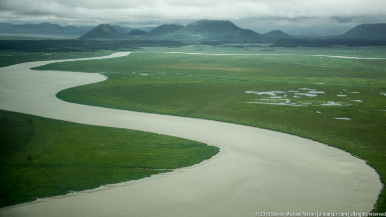Aerial view of a pale winding river crossing green wetlands, with forest and mountains beneath low clouds.