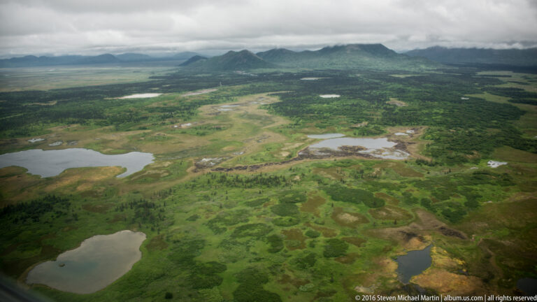 Aerial view across a green wetland plain dotted with ponds, with a low mountain ridge under gray clouds.