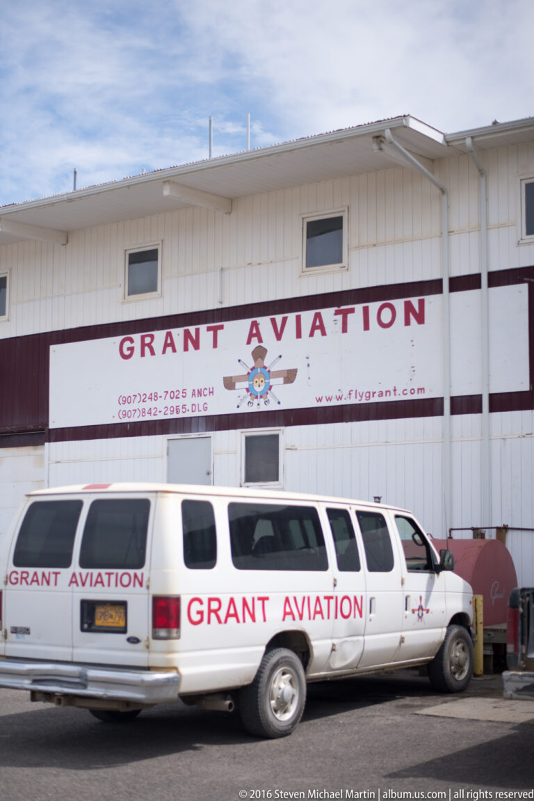 White Grant Aviation passenger van parked beside a matching company sign on a metal-sided building.