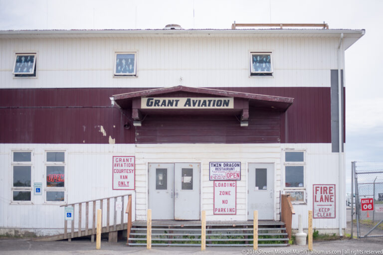 White-and-burgundy Grant Aviation building with a covered entrance, accessibility ramp, posted notices, and marked fire lane.