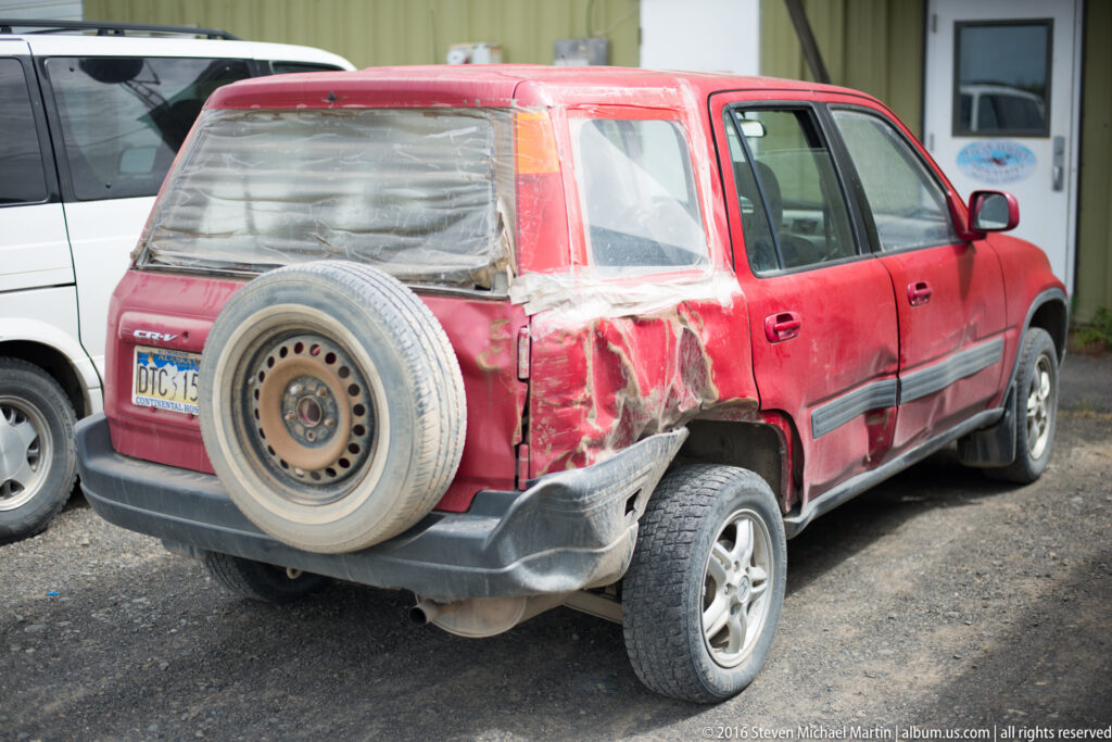 Red SUV parked on gravel, with a spare tire on the rear door and deep damage to the left rear quarter.