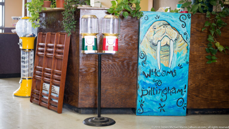 Turquoise hand-painted sign reading “Welcome to Dillingham,” with a fish, beside chairs and beverage dispensers.