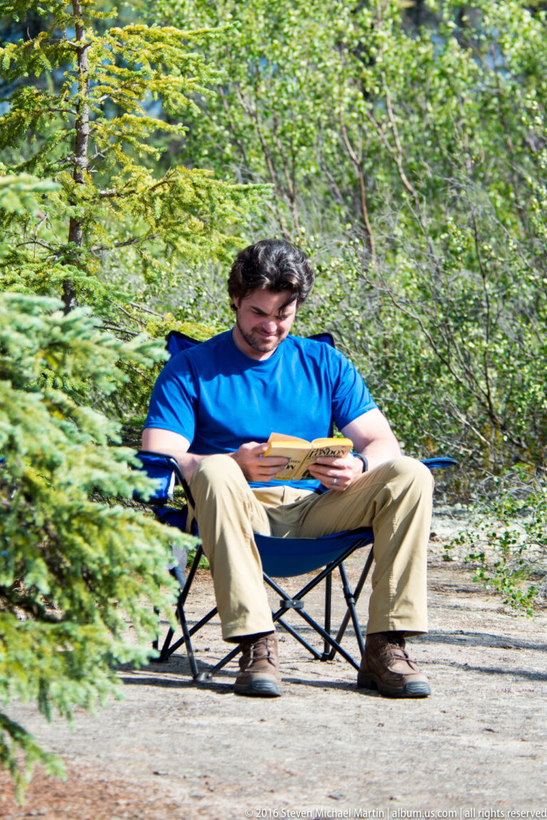 A man in a blue shirt sits on a folding chair on a gravel track and reads a small booklet.