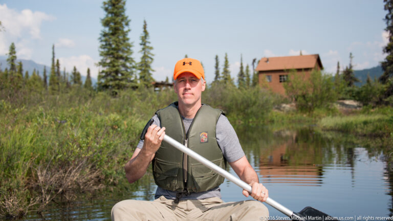 A man in an orange cap and flotation vest sits in a canoe with a paddle on a calm, shrub-lined lake.