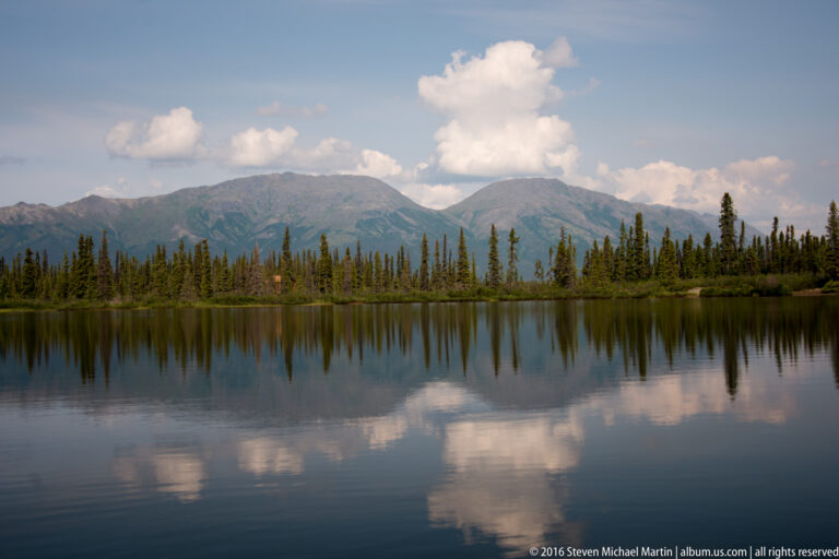 A tall white cloud and mountain ridge reflect in a calm lake beyond a band of spruce trees.