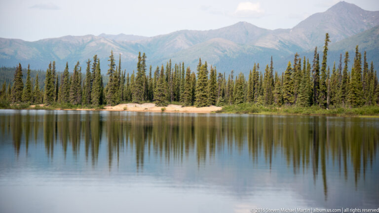 A spruce-covered sandy point extends into a still lake, with a mountain slope and shoreline reflected in the water.