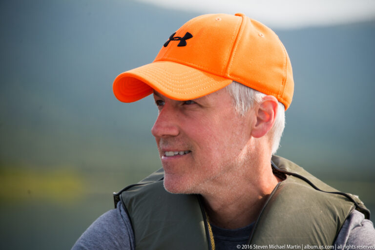 A man in an orange cap and green flotation vest looks left against a softly blurred background.